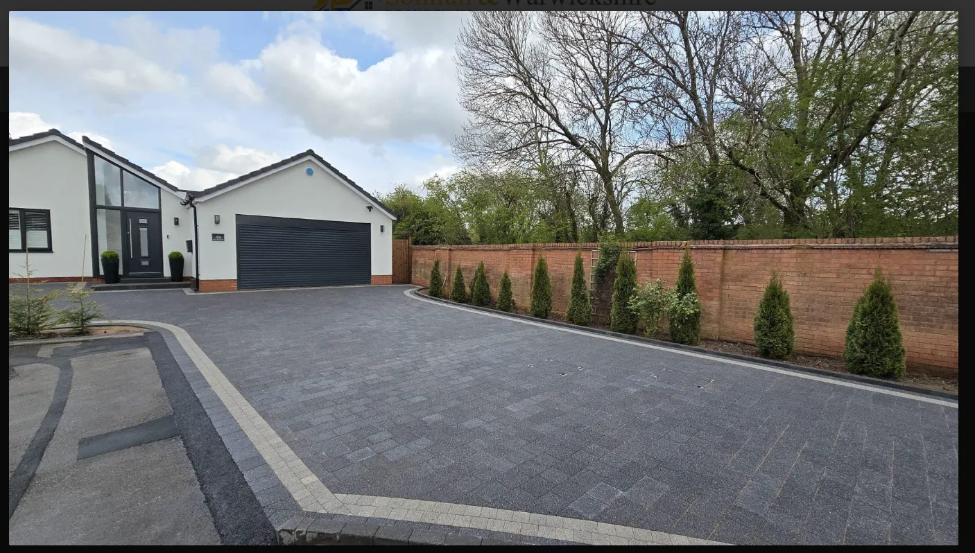 Modern white house with a dark garage door, surrounded by neatly trimmed bushes and a brick wall, under a partly cloudy sky.