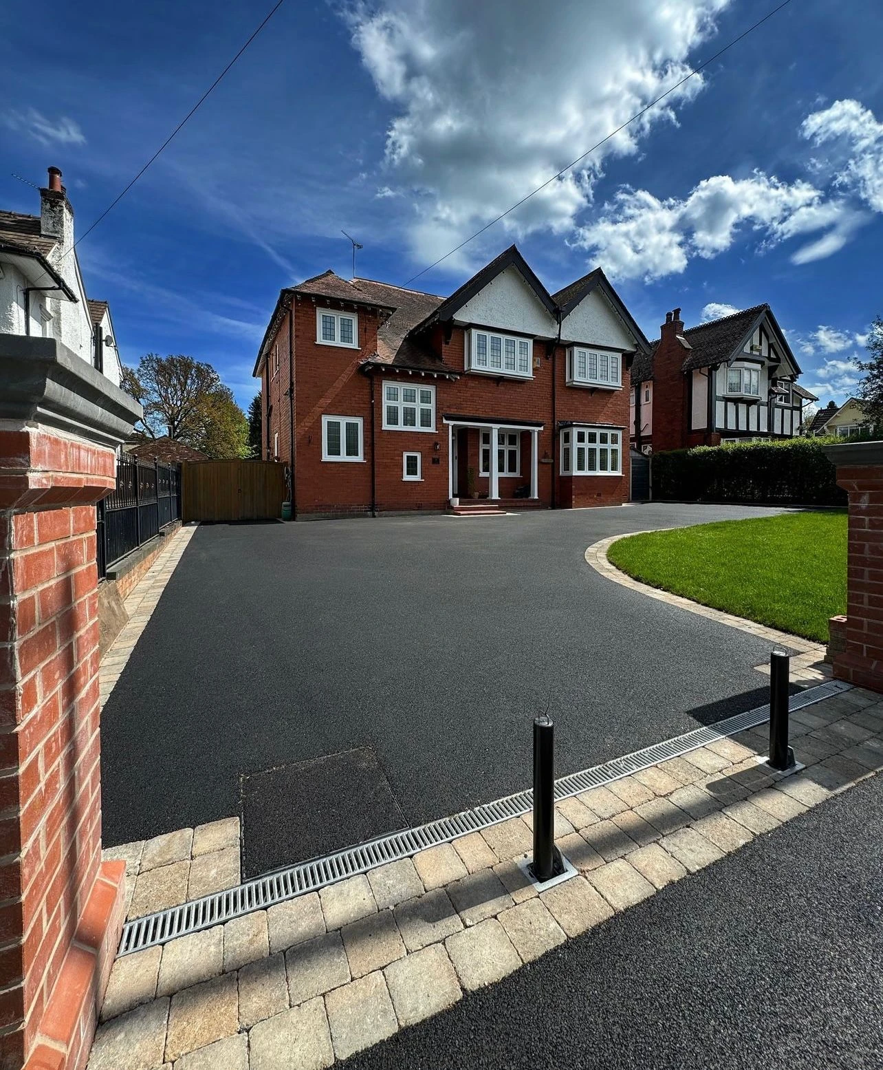 A large red-brick house with a manicured lawn and paved driveway under a blue sky with clouds. A modern fence encloses the property.