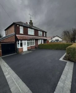 Two-story brick house with hedges and a freshly paved driveway under a cloudy sky.