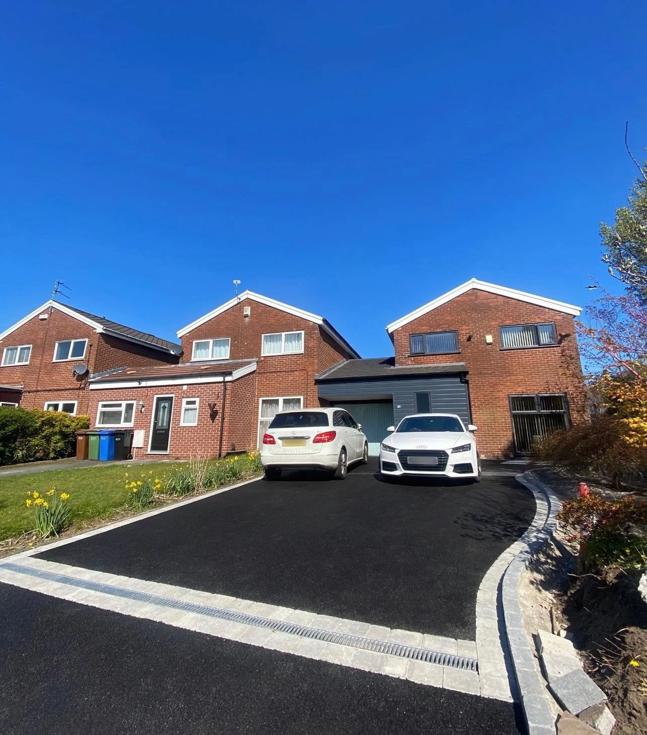 Two brick houses with a black driveway, featuring a white car and a silver car parked outside, under a clear blue sky.