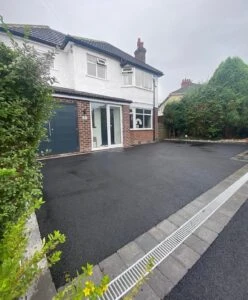 A modern two-story house with a dark driveway and greenery nearby, featuring a large front window and a garage.