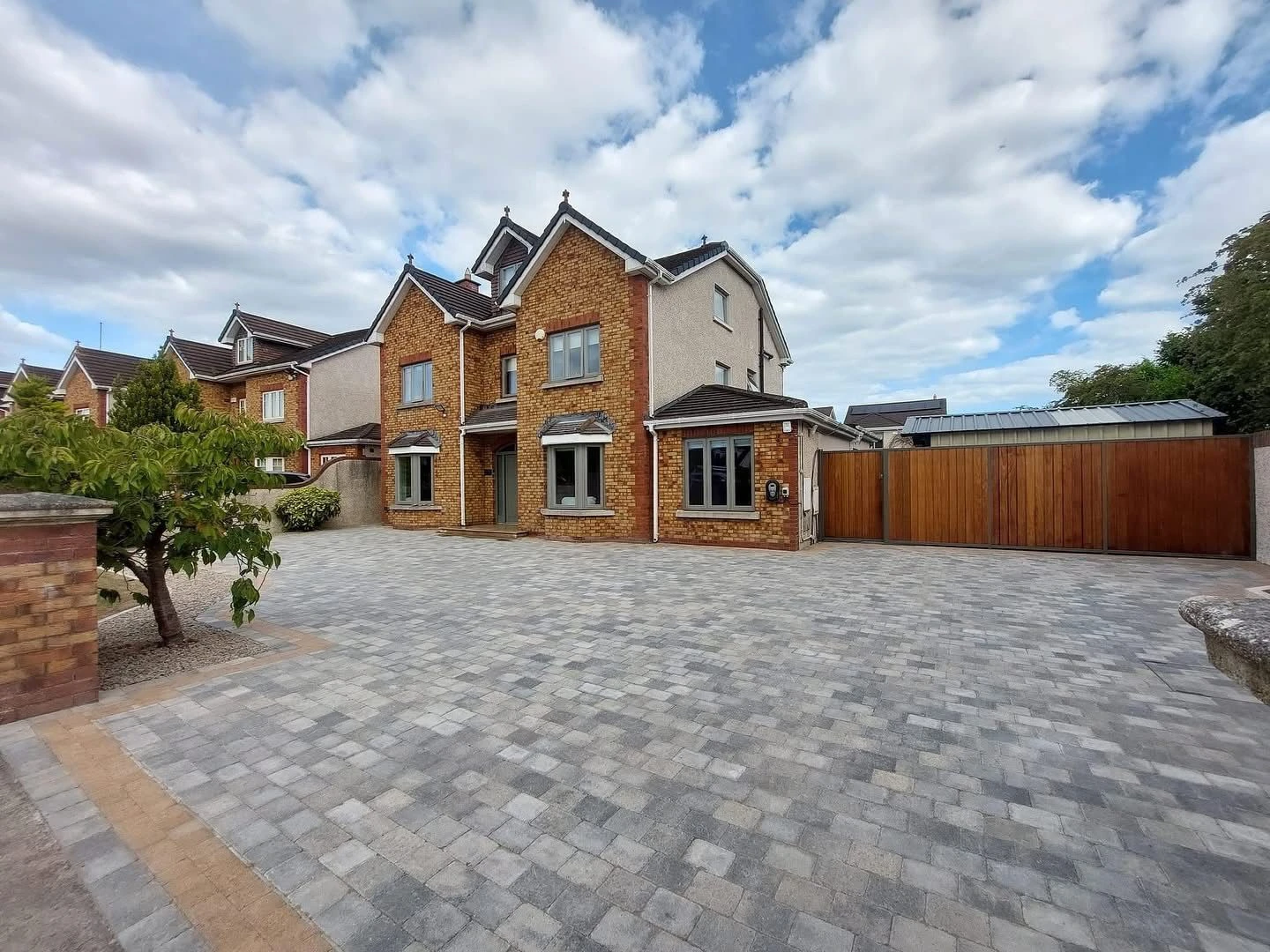 A spacious two-story brick house with a paved driveway and wooden gate, surrounded by greenery and a cloudy sky.