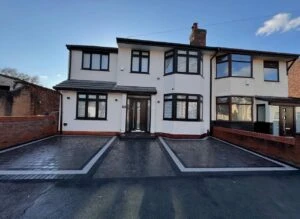 Modern two-story house with a symmetrical facade, large windows, and a paved driveway. Clear blue sky in the background.