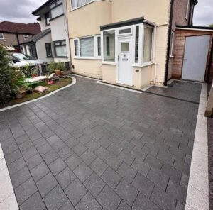 Residential driveway with gray pavers, leading to a house with a white door and a side shed, surrounded by greenery.