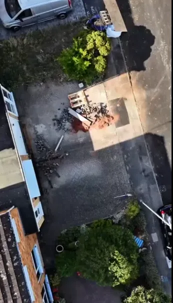 Aerial view of a residential area with construction debris on a cobblestone driveway, surrounded by greenery and parked vehicles.