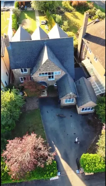 Aerial view of a large house with a stone facade, surrounded by greenery and a driveway, showcasing a well-maintained garden.